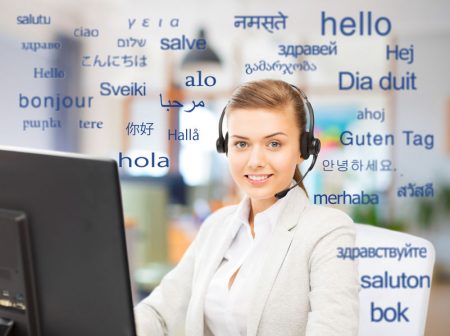 translation, business and technology concept - smiling female translator or operator with computer at office over greeting words in different foreign languages