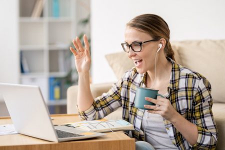 Cheerful excited young woman in earphones sitting at home and waving hand while talking to colleagues via Internet